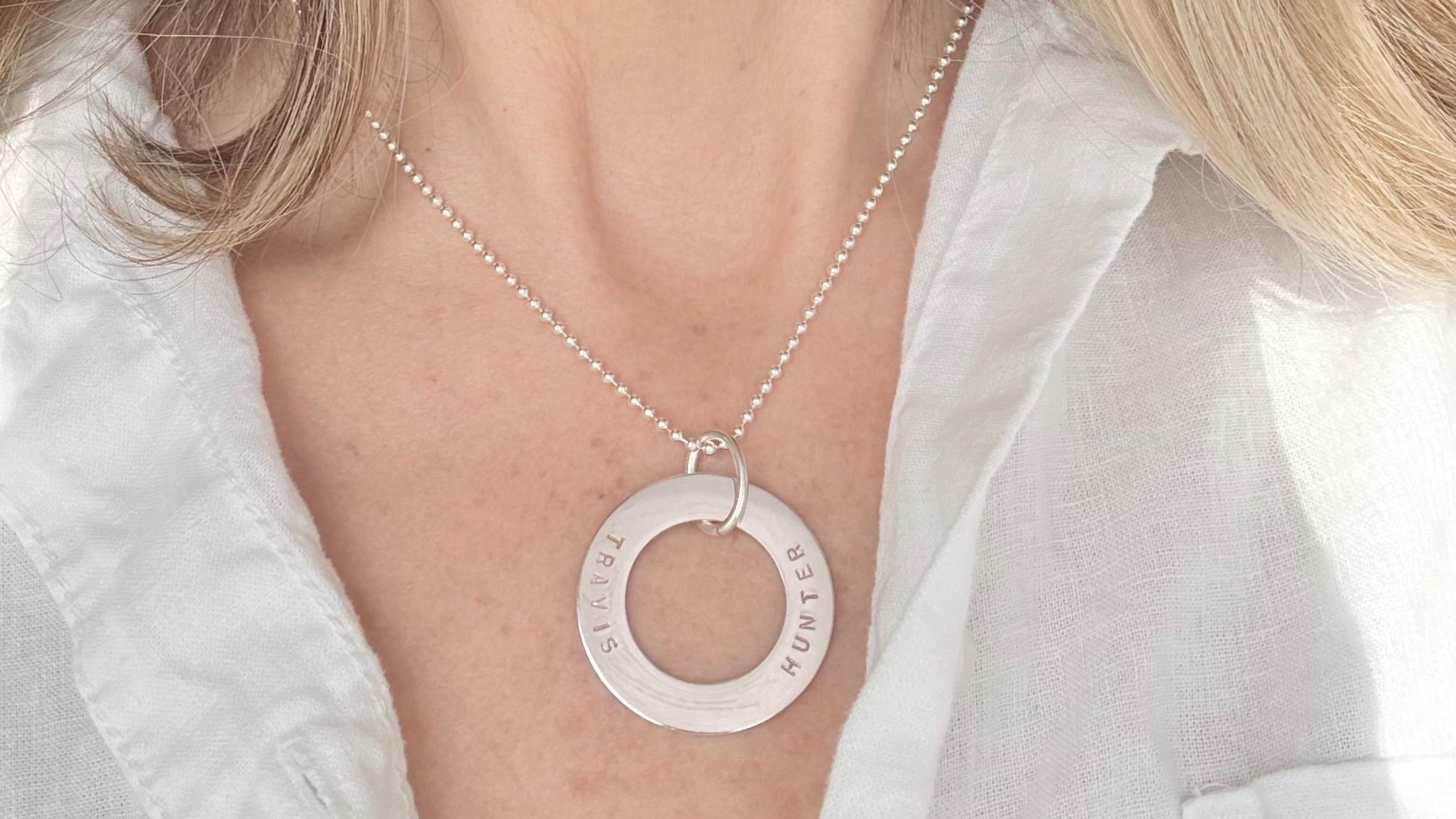 Person wearing a silver necklace with a circular pendant against a neutral background