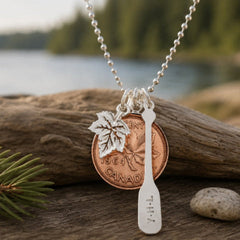 Necklace with a Canadian penny and leaf charm on a wooden log by a lake.