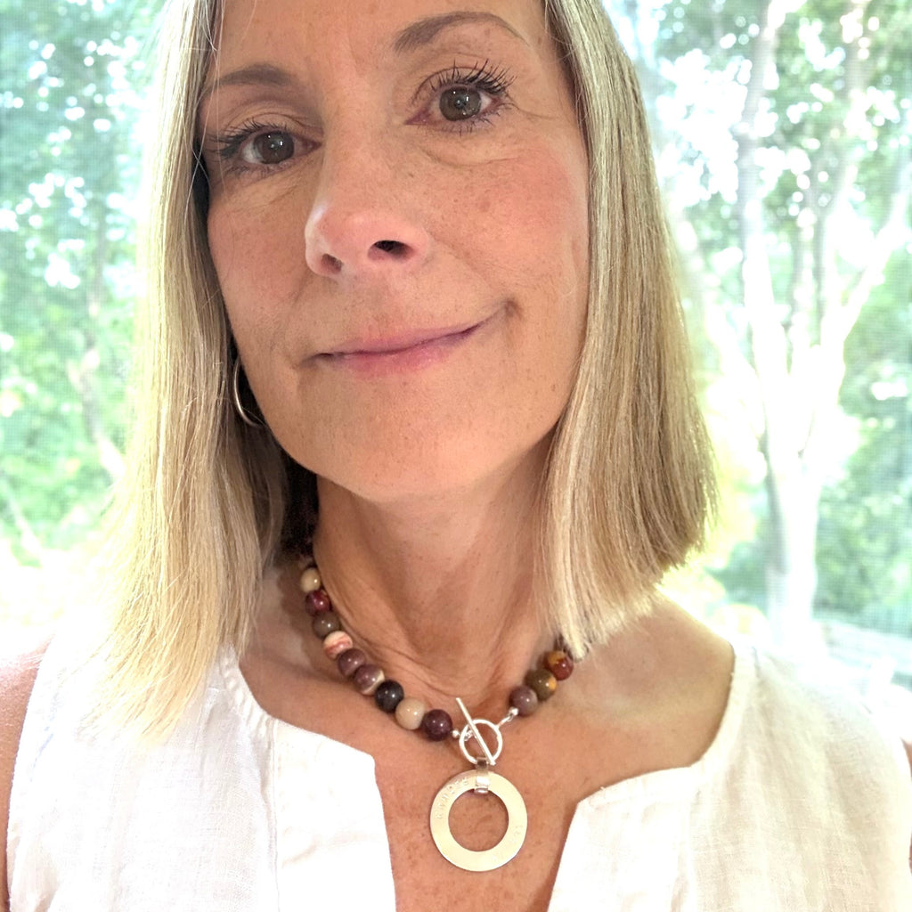 Woman wearing a necklace with a large circular pendant in front of a window with greenery.