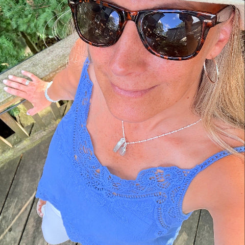 A woman wearing a blue eyelet camisole top, tortoiseshell sunglasses, a white hat, and a silver pendant necklace, taking a selfie on a wooden deck.