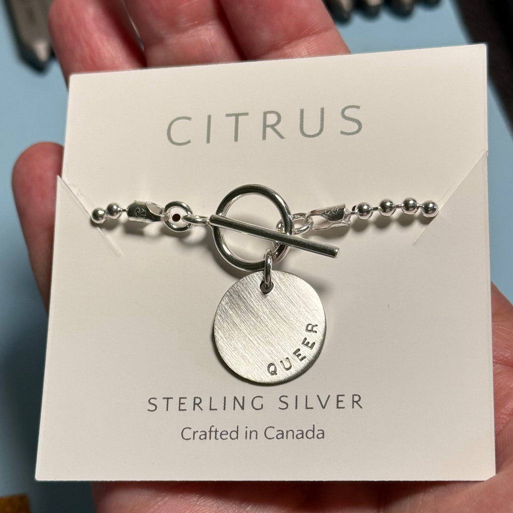 Silver chain with silver pendant hand stamped with QUEER on a 'Citrus' branded card held by a hand.