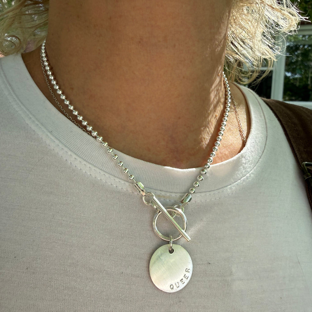 Close-up of a silver Love Necklace with a round pendant hand stamped with QUEER by CITRUS