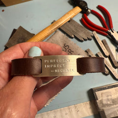 Hand holding a leather tag with 'Perfectly Imperfect Michael' branding against a workshop background.