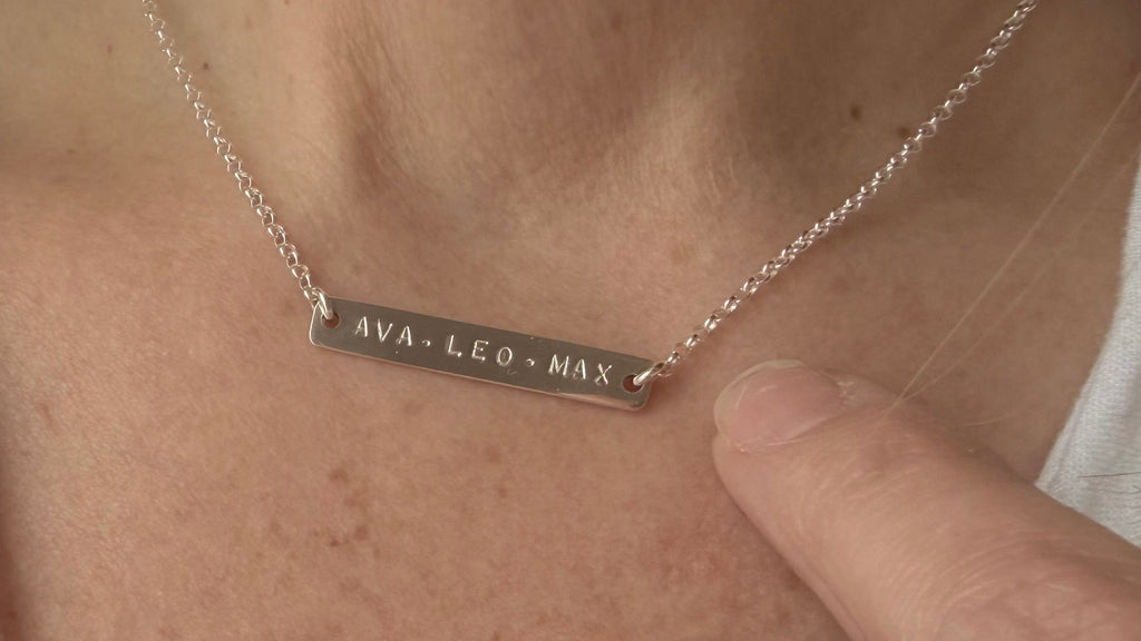 Necklace with engraved name bar worn by a person, close-up of neck and hand.