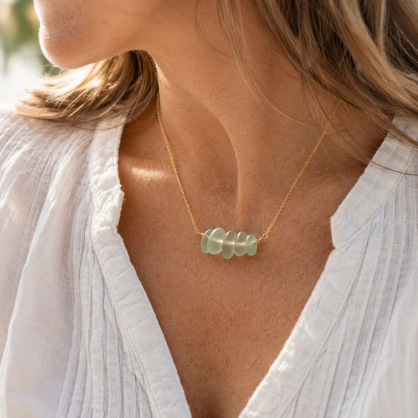 A close-up lifestyle shot of a woman wearing a delicate gold chain necklace with four smooth, pale green stone beads at the center, styled with a white textured blouse.