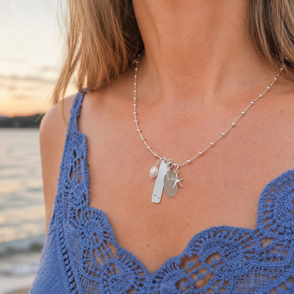 A close-up of a woman wearing a silver beaded necklace with a lighthouse and starfish pendant, paired with a blue crochet top, with a blurred beach sunset background.