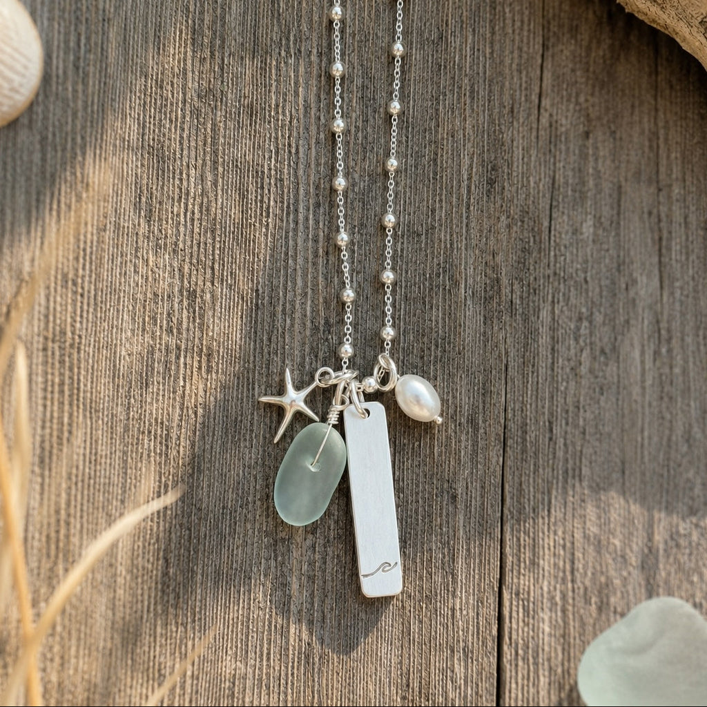 A silver charm necklace with a beaded chain, featuring a starfish, a baroque shell, a small pearl, and a rectangular engraved plate, displayed on a rustic wooden surface with dried grass and seashell props.
