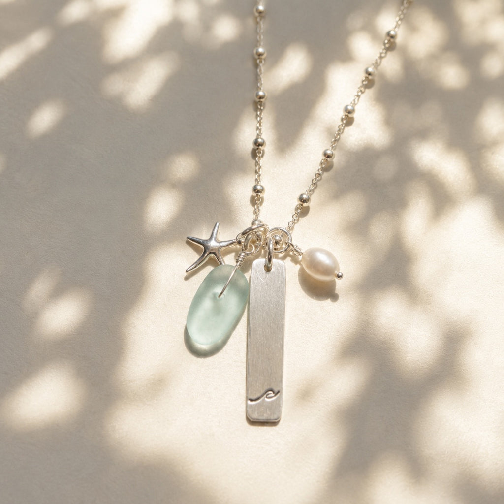 A delicate silver beaded necklace with a vertical bar pendant, a starfish charm, a light green sea glass piece, and a small white pearl charm, displayed against a warm beige background with soft dappled shadows.