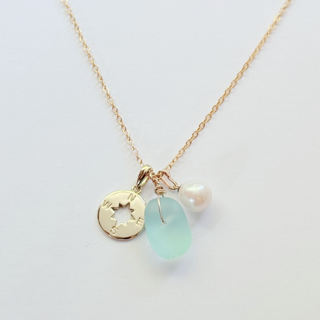 A delicate gold chain necklace featuring three charms: a gold star-cut coin pendant, a light blue sea glass bead, and a small white pearl, displayed on a clean white background.
