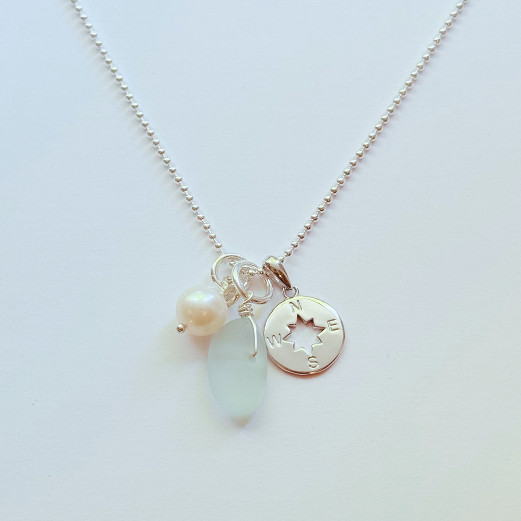 A delicate silver ball-chain necklace with a cluster of charms, including a small white pearl bead, a smooth pale blue teardrop stone, and a round silver compass coin charm.