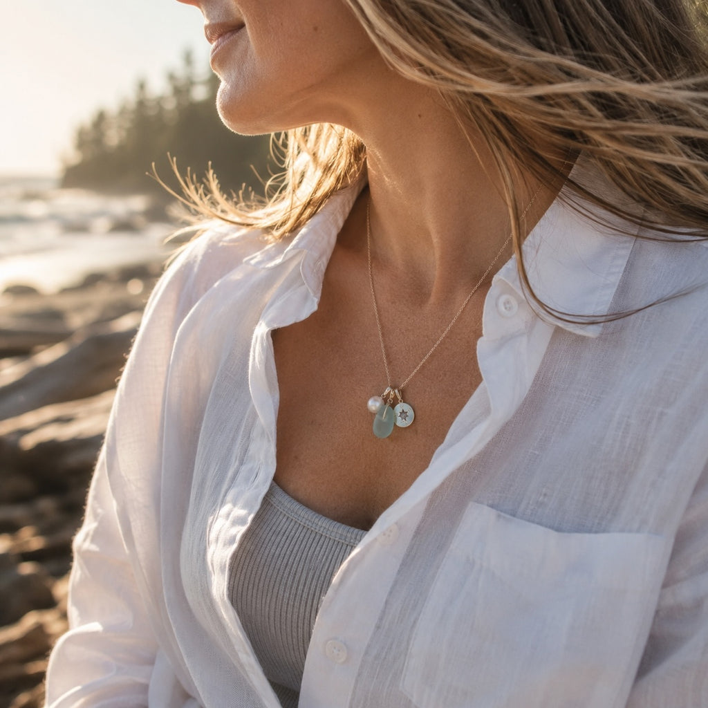 Woman sitting on a beach wearing a golden necklace with a compass, a freshwater pearl and sea glass