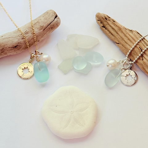 Two delicate beach-themed necklaces displayed on driftwood branches with sea glass and a sand dollar shell. The left necklace is gold with a gold coin charm, a white pearl, and a light blue sea glass bead.