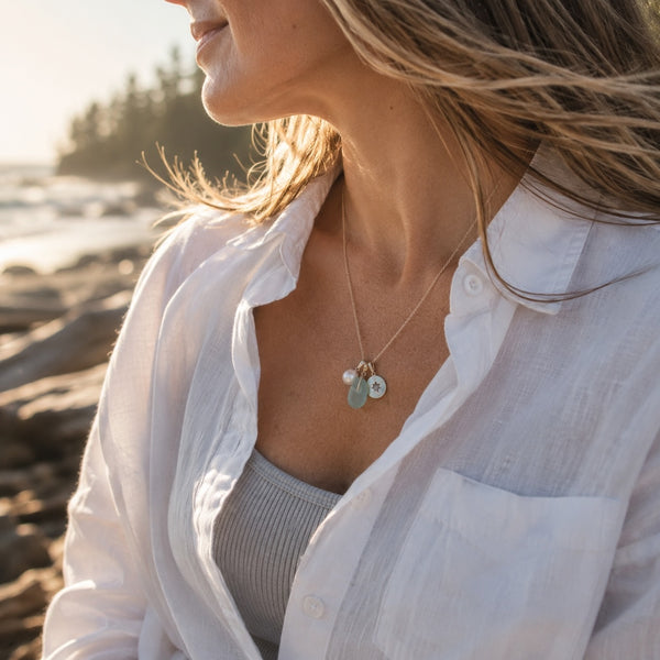 Woman sitting on a beach wearing a golden necklace with a compass, a freshwater pearl and sea glass